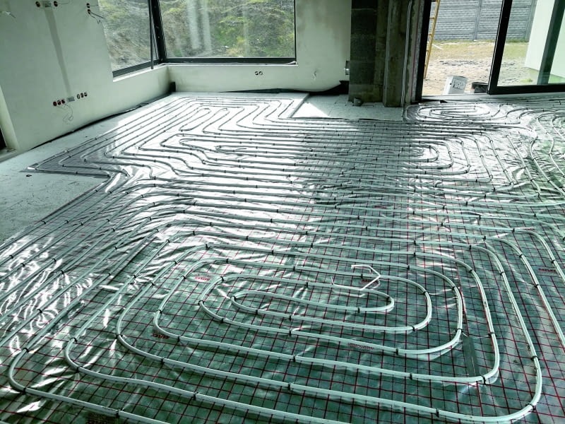 Rozłożona instalacja ogrzewania podłogowego na folii z siatką w surowym wnętrzu. Widoczne rury grzewcze w kształcie meandrów i pętelek przed wylaniem posadzki.