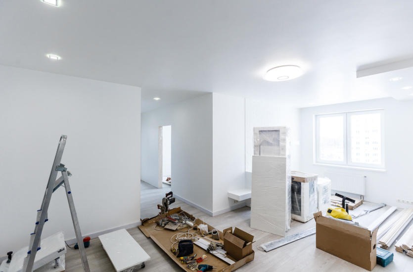 Jasne wnętrze w trakcie remontu: drabina, narzędzia, materiały budowlane, okno z widokiem na zewnątrz. Widoczne ślady świeżego malowania ścian.
