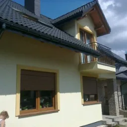 Jasny dom jednorodzinny z ciemną dachówką, widoczne okna z roletami, balkon z ozdobną balustradą i elementy wykończenia elewacji z kamienia.