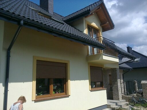Jasny dom jednorodzinny z ciemną dachówką, widoczne okna z roletami, balkon z ozdobną balustradą i elementy wykończenia elewacji z kamienia.