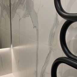 MOORE Karol Derbin - Białe płytki imitujące marmur na ścianie łazienki z czarnym, designerskim grzejnikiem drabinkowym i fragmentem lustra z podświetleniem LED.