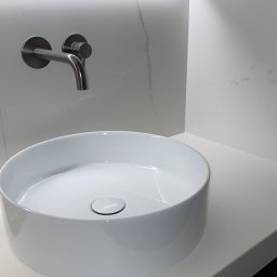 MOORE Karol Derbin - Biała, okrągła umywalka nablatowa z chromowaną baterią na tle marmurowej ściany z podświetleniem LED. Minimalistyczny design, nowoczesne wykończenie łazienki.