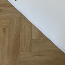 MOORE Karol Derbin - Ukończona podłoga z paneli ułożonych w jodełkę, kontrastująca z białą ścianą i widocznym fragmentem listwy przypodłogowej. Minimalistyczne ujęcie.