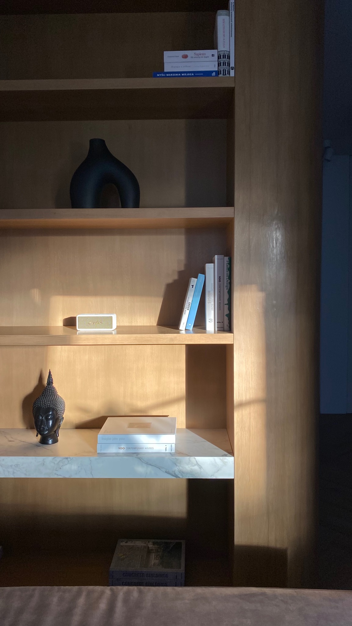 Drewniany regał z dekoracjami: wazon, głośnik, książki i głowa Buddy na marmurowej półce. Ciepłe światło podkreśla fakturę drewna.