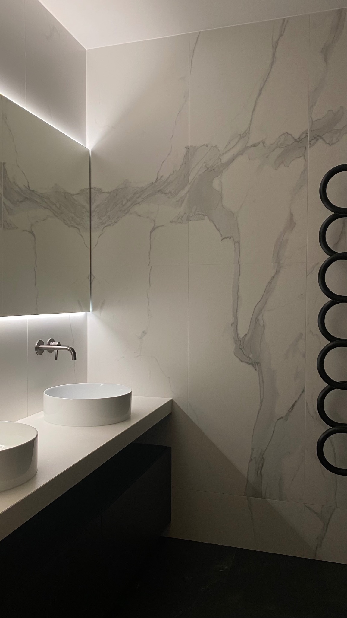 Nowoczesna łazienka z marmurowymi płytkami, podświetlanym lustrem i okrągłymi umywalkami nablatowymi. Minimalistyczny design z czarnymi akcentami.