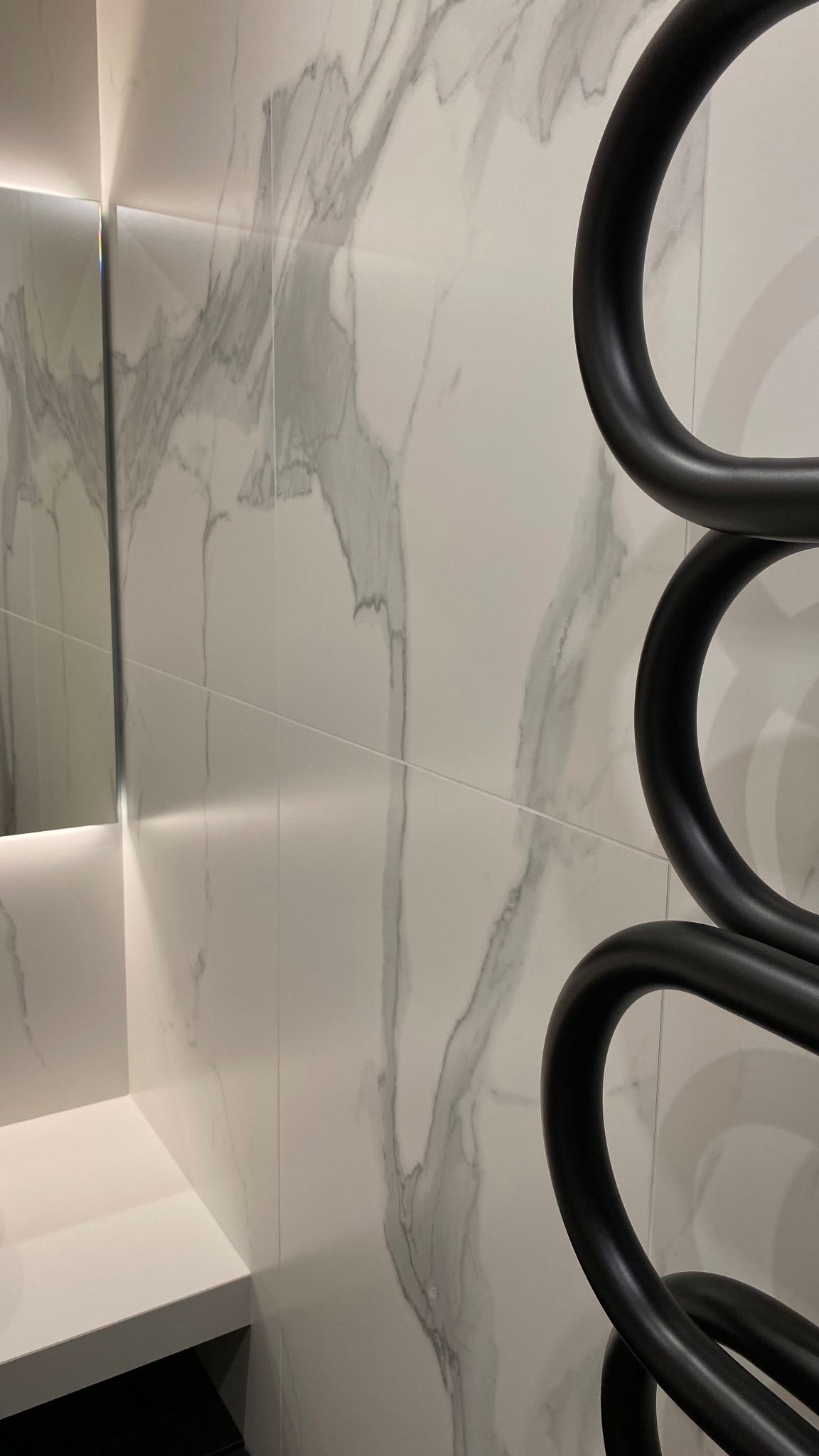 Białe płytki imitujące marmur na ścianie łazienki z czarnym, designerskim grzejnikiem drabinkowym i fragmentem lustra z podświetleniem LED.