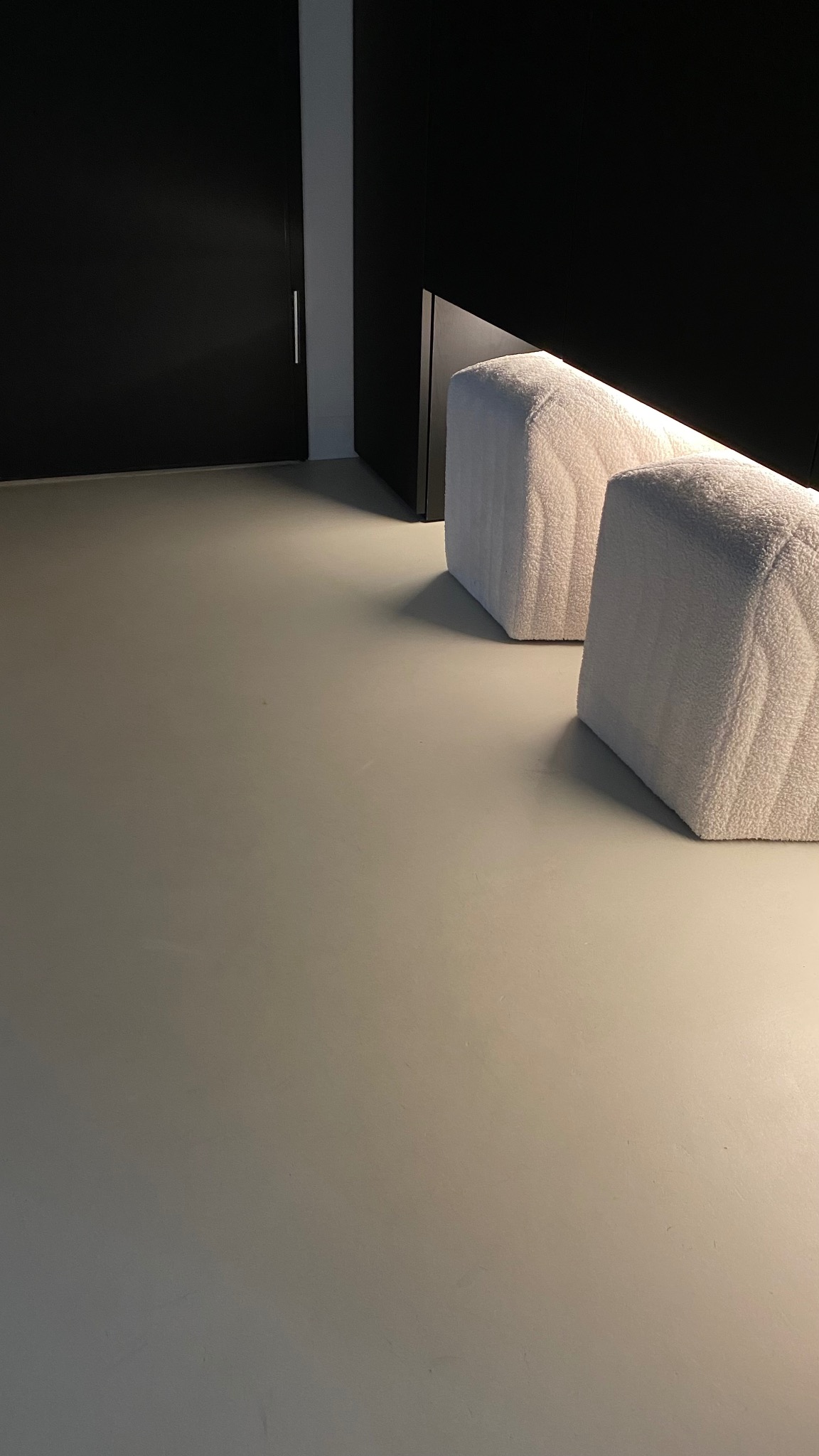 Minimalistyczne wnętrze: gładka, szara podłoga, dwa białe pufy o fakturze baranka podświetlone od dołu, czarne szafy w tle. Nowoczesny design.