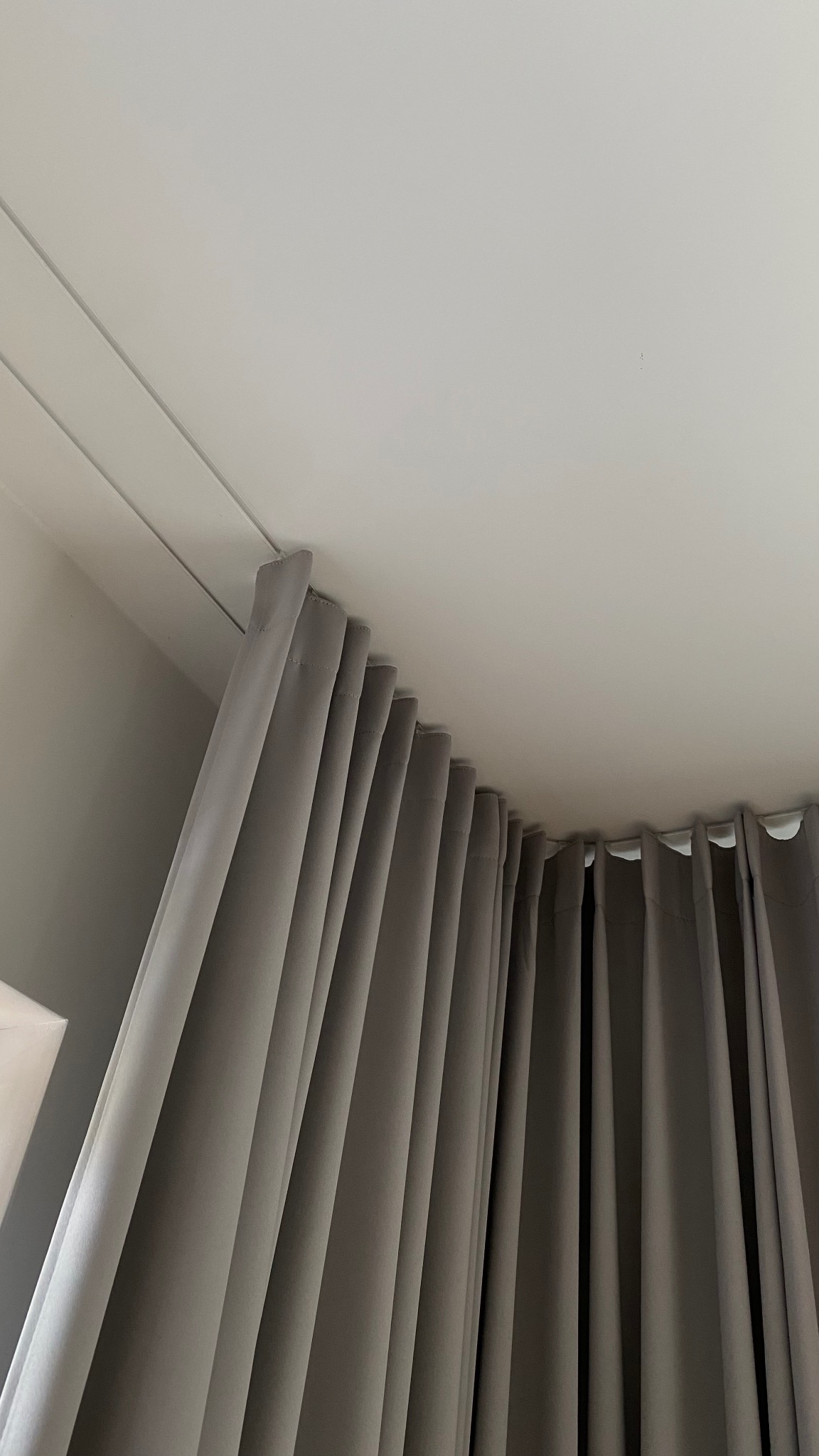 Szare, gładkie zasłony wiszące na karniszu zamontowanym pod sufitem. Ujęcie od dołu, widoczny fragment sufitu i górna część zasłon z równomiernymi fałdami.