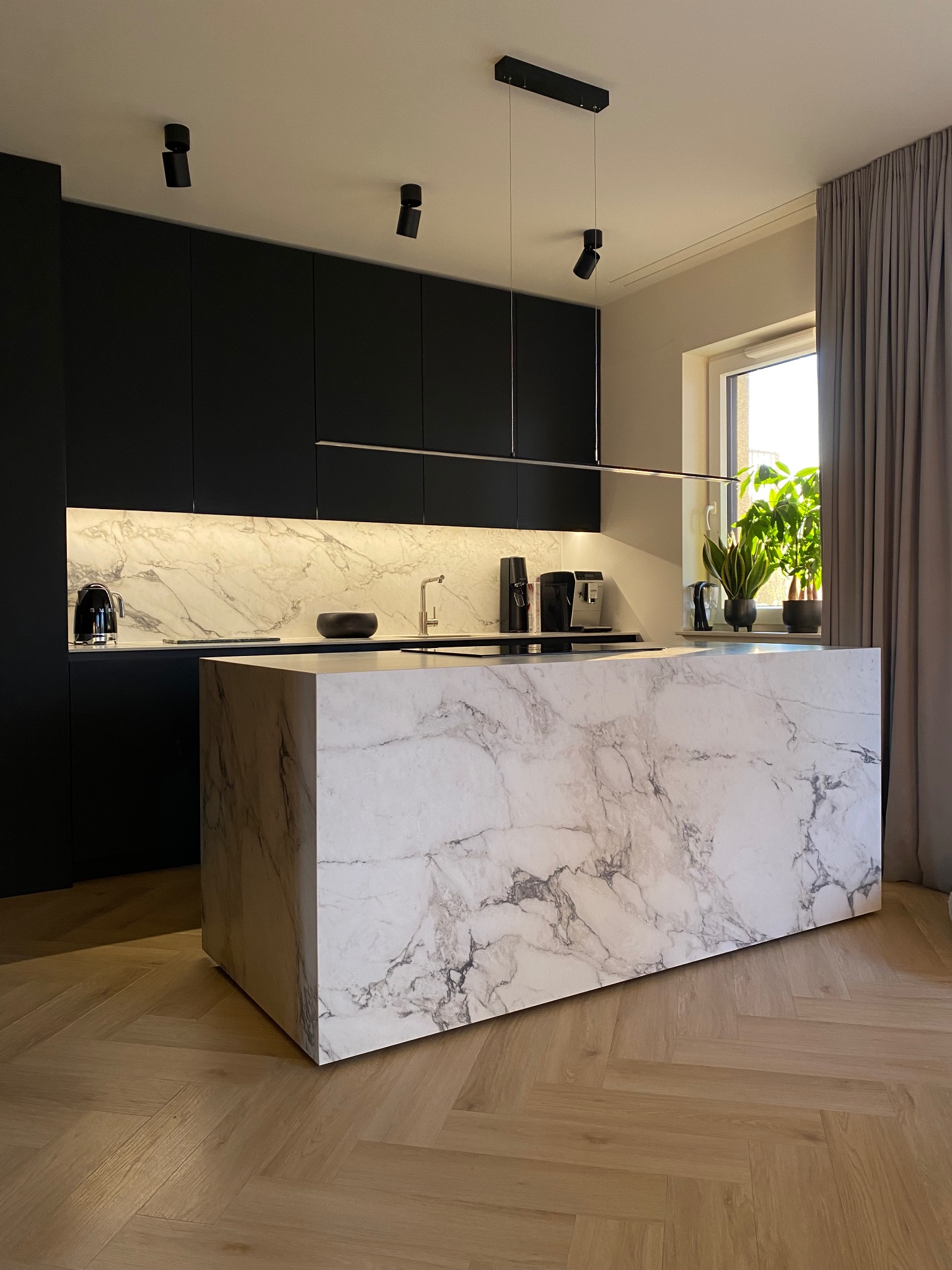Nowoczesna kuchnia z wyspą wykończoną marmurem, czarne szafki, parkiet w jodełkę, oświetlenie punktowe i okno z roślinami. Minimalistyczny design.