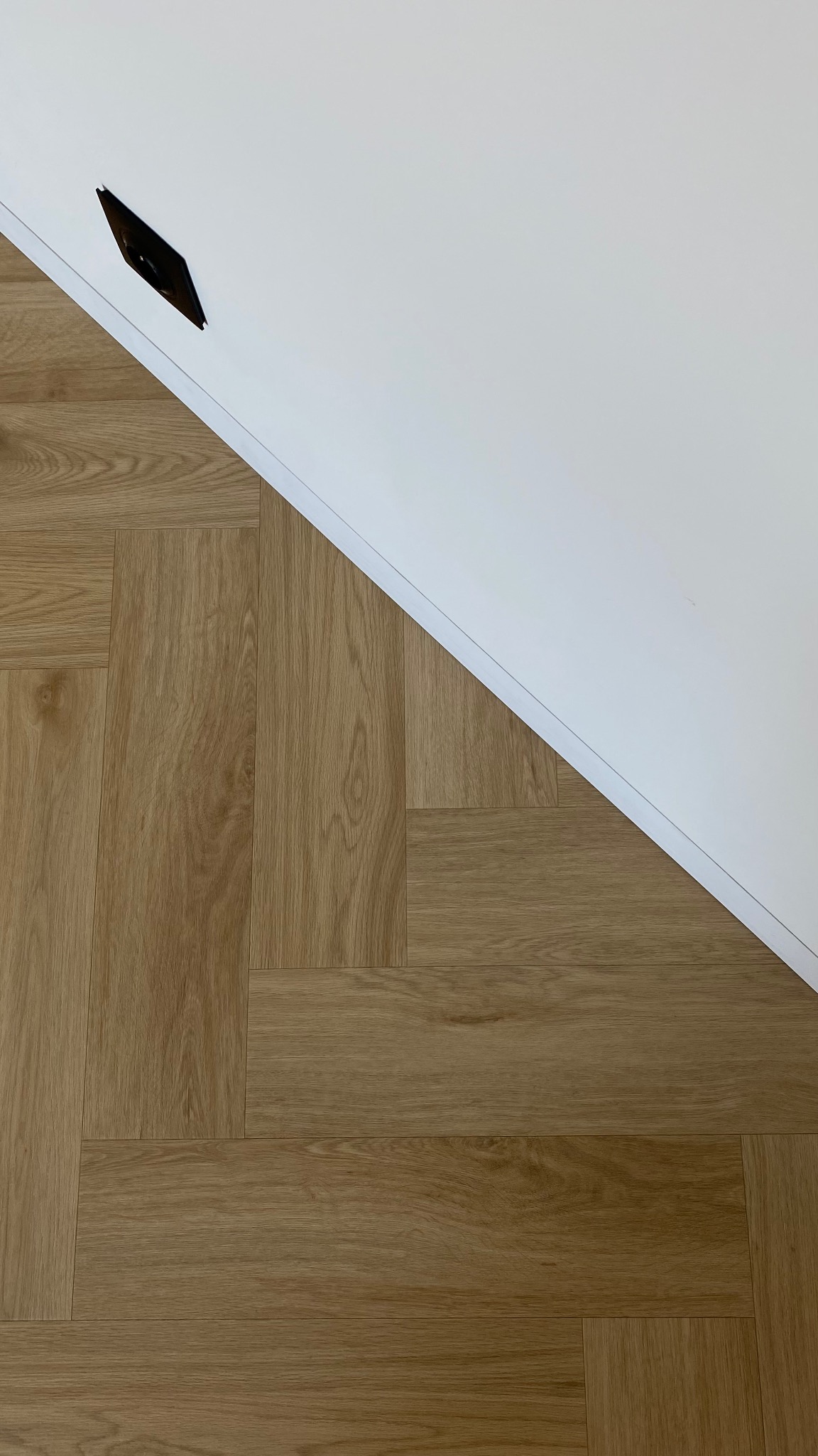 Ukończona podłoga z paneli ułożonych w jodełkę, kontrastująca z białą ścianą i widocznym fragmentem listwy przypodłogowej. Minimalistyczne ujęcie.