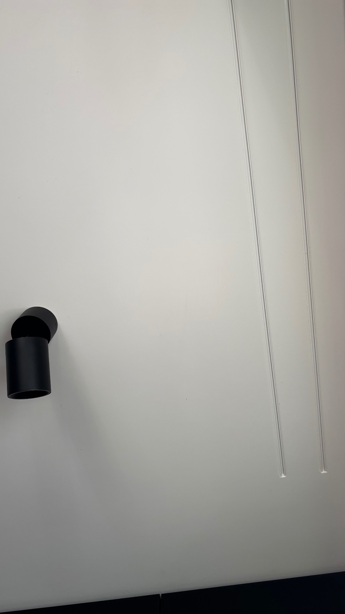 Minimalistyczna, biała ściana z dwoma pionowymi listwami i nowoczesnym, czarnym kinkietem. Kinkiet składa się z dwóch cylindrycznych elementów.