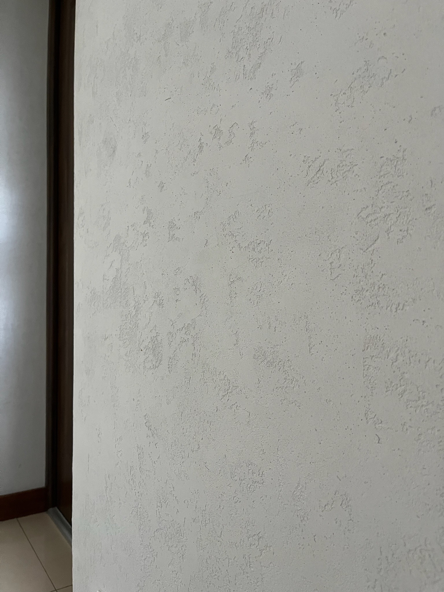 Ściana z dekoracyjnym, jasnym tynkiem strukturalnym, widoczna z bliska, z fragmentem ościeżnicy drzwi po lewej stronie. Delikatna faktura i subtelne cieniowanie.