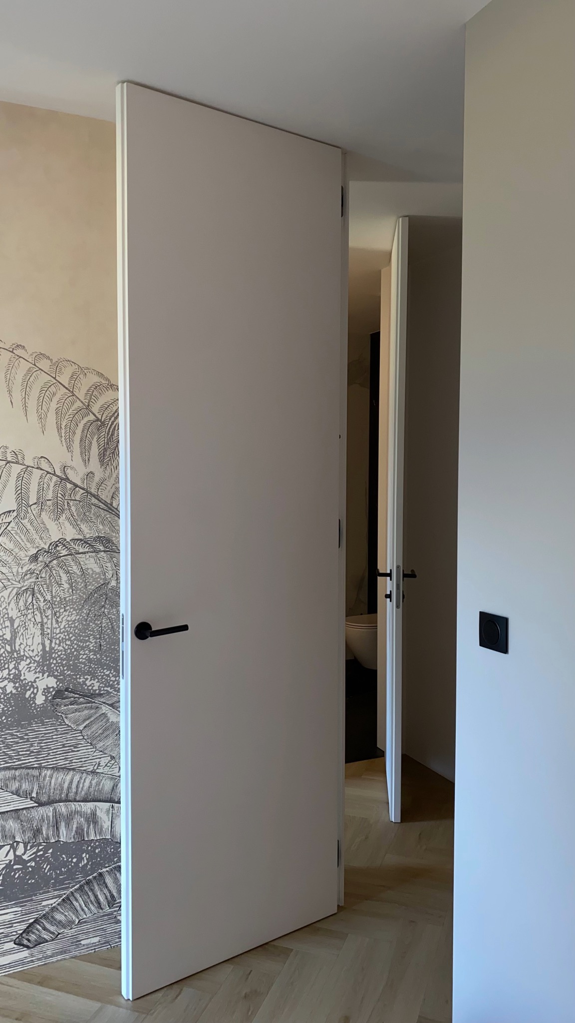 Białe, minimalistyczne drzwi z czarną klamką, otwarte do wnętrza z widoczną toaletą. Na ścianie tapeta z motywem roślinnym i nowoczesny włącznik.