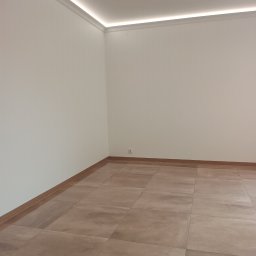 URB MZ - Jasne, beżowe płytki podłogowe w nowym pokoju z białymi ścianami i listwami sufitowymi z oświetleniem LED. Widoczny fragment gniazdka elektrycznego.