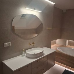 URB MZ - Nowoczesna łazienka z owalnym lustrem, białą umywalką nablatową i złotą armaturą. Podświetlenie LED podkreśla minimalistyczny design i ciepłą kolorystykę płytek.
