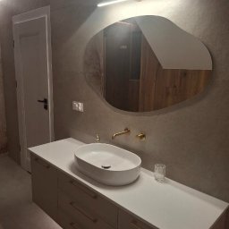 URB MZ - Nowoczesna łazienka z owalną umywalką nablatową, złotą armaturą i asymetrycznym lustrem podświetlonym LED. Minimalistyczny design, szare płytki i białe meble.