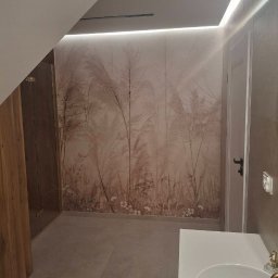 URB MZ - Wykończona łazienka na poddaszu ze ścianą w beżowe trawy, oświetleniem LED i umywalką. Widoczny fragment kabiny prysznicowej i drzwi.