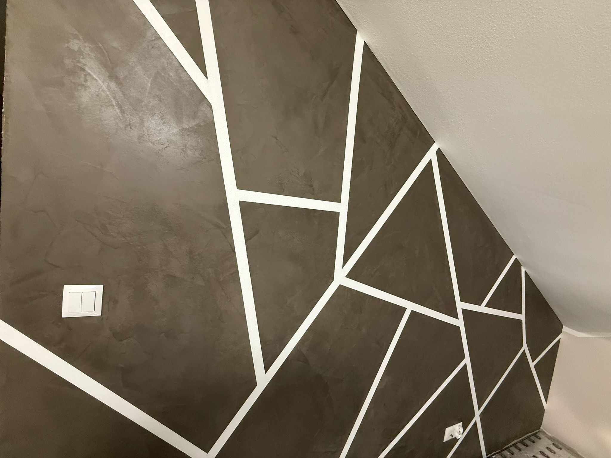 Ściana w odcieniach brązu z geometrycznym wzorem z białych pasów, widoczny włącznik światła i fragment sufitu. Dekoracyjne wykończenie wnętrza.