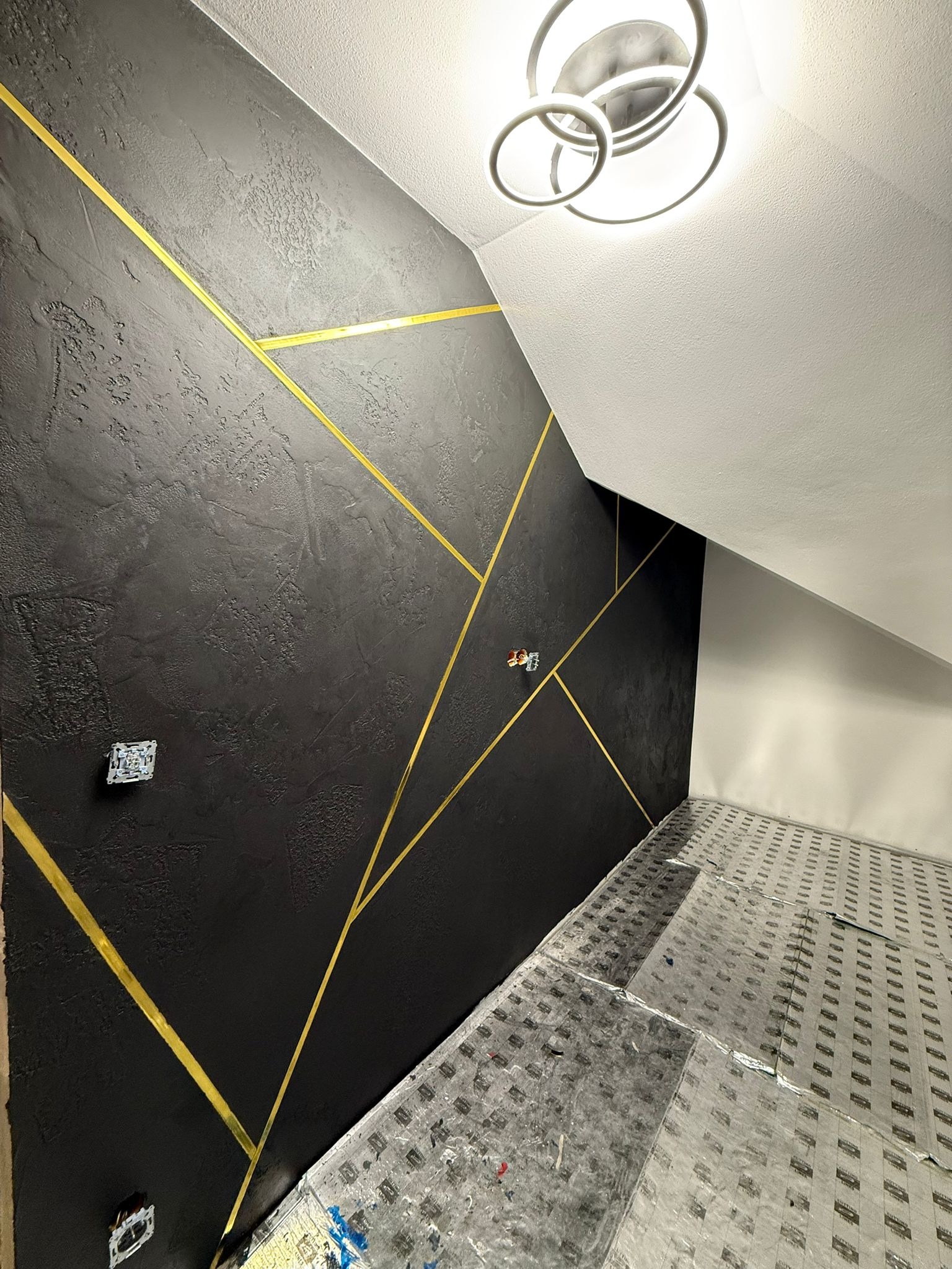 Czarna ściana z dekoracyjną strukturą i złotymi paskami w skośnym układzie, widoczne gniazdka elektryczne, sufit ze skośnym załamaniem i nowoczesna lampa.