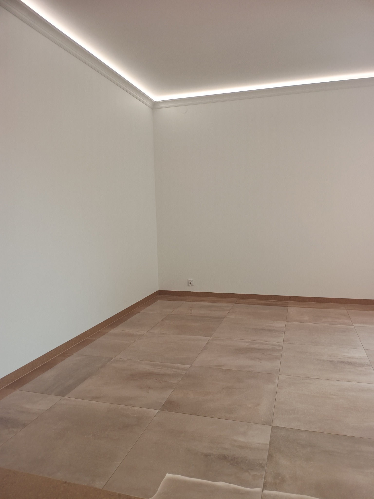 Jasne, beżowe płytki podłogowe w nowym pokoju z białymi ścianami i listwami sufitowymi z oświetleniem LED. Widoczny fragment gniazdka elektrycznego.