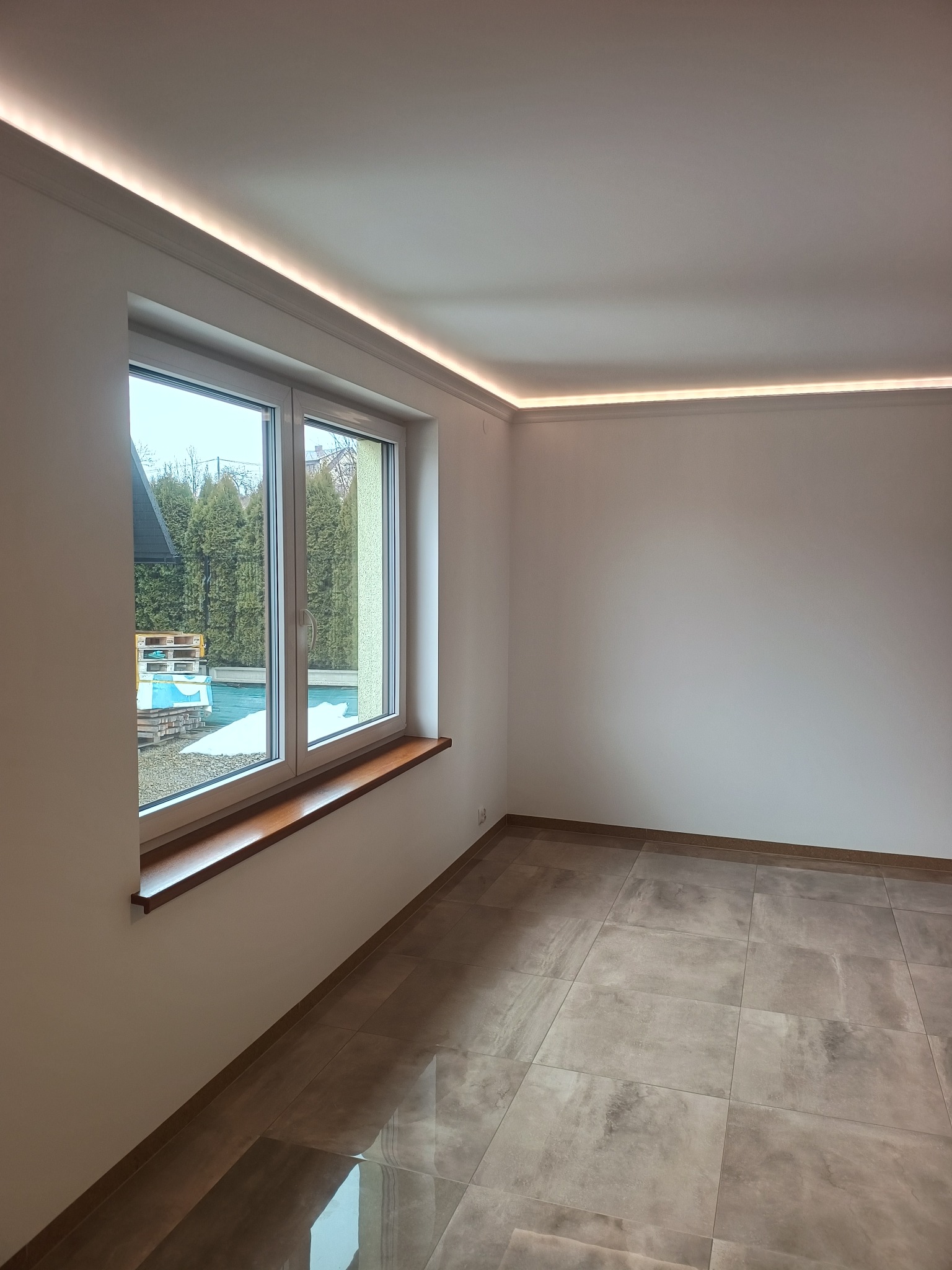 Jasne wnętrze po remoncie: brązowe płytki podłogowe, białe ściany, okno z widokiem na zieleń i subtelne oświetlenie LED podsufitowe. Minimalistyczny design.