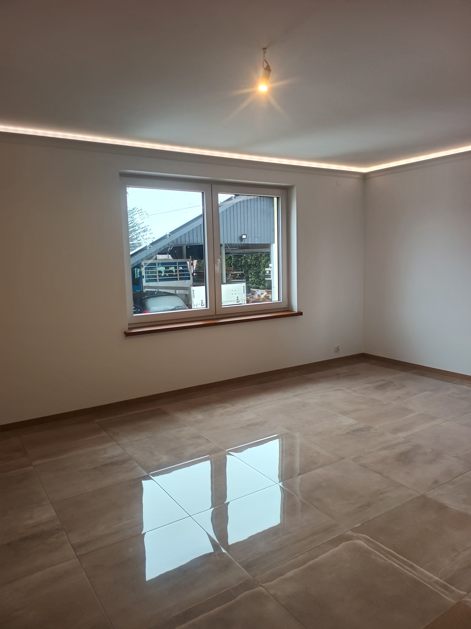 Wykończone wnętrze pokoju z błyszczącą, beżową podłogą z płytek, białymi ścianami, oknem i subtelnym oświetleniem LED na suficie. Widok na zewnątrz przez okno.