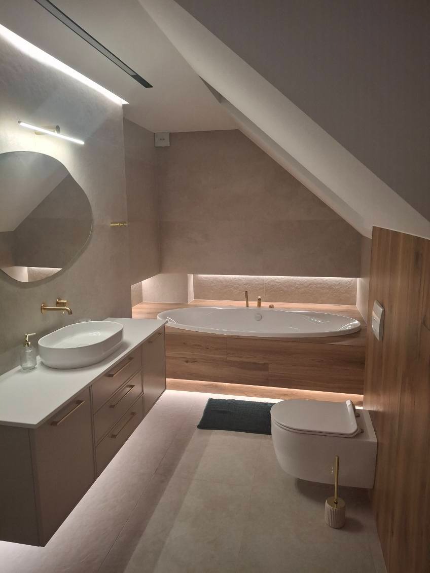 Nowoczesna łazienka na poddaszu z owalną wanną, umywalką nablatową i podświetleniem LED. Uwagę zwraca nietypowe lustro o organicznym kształcie i złote akcenty.