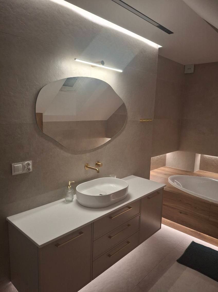 Nowoczesna łazienka z owalnym lustrem, białą umywalką nablatową i złotą armaturą. Podświetlenie LED podkreśla minimalistyczny design i ciepłą kolorystykę płytek.