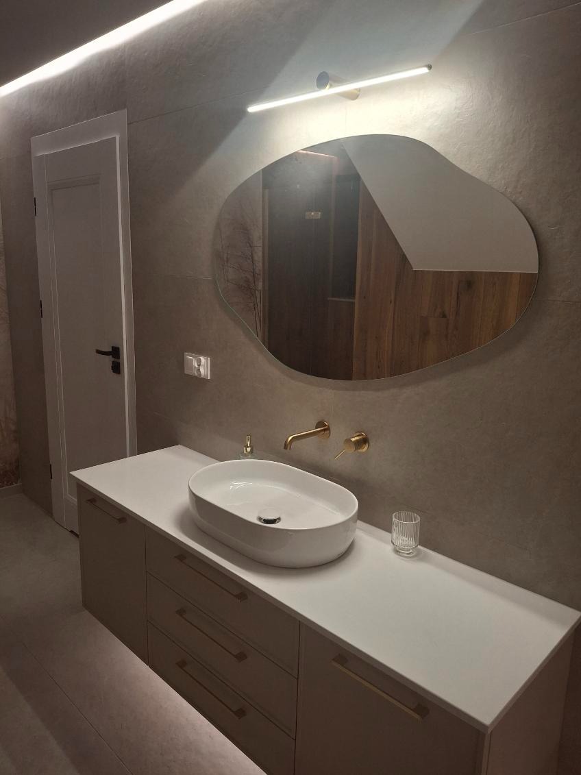 Nowoczesna łazienka z owalną umywalką nablatową, złotą armaturą i asymetrycznym lustrem podświetlonym LED. Minimalistyczny design, szare płytki i białe meble.