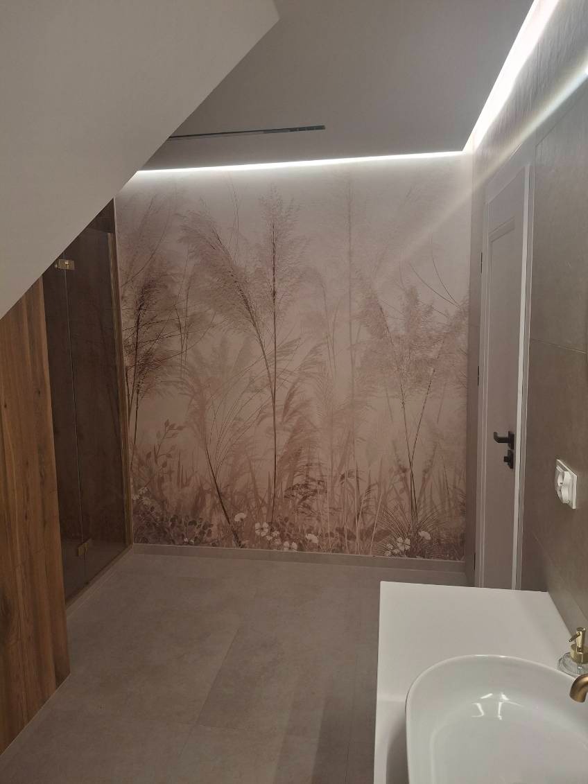 Wykończona łazienka na poddaszu ze ścianą w beżowe trawy, oświetleniem LED i umywalką. Widoczny fragment kabiny prysznicowej i drzwi.