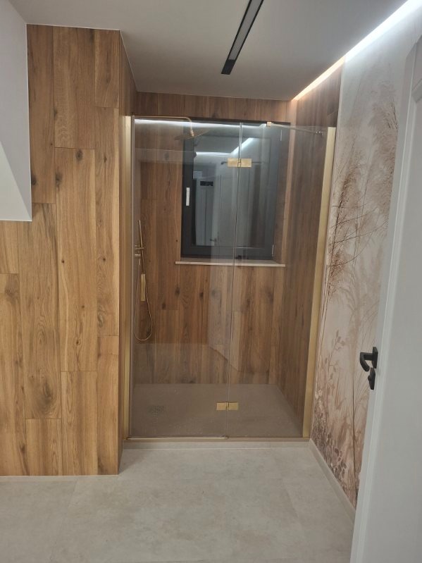 Nowoczesna kabina prysznicowa z drewnopodobnymi płytkami na ścianach i złotymi detalami, oświetlona ledami, z oknem wewnątrz kabiny i dekoracyjną tapetą na ścianie.