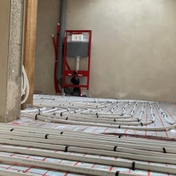 AQUA HORIZON SPÓŁKA Z OGRANICZONĄ ODPOWIEDZIALNOŚCIĄ - Białe rury ogrzewania podłogowego ułożone na czerwonej macie w surowym wnętrzu, widok przez framugę drzwi, z elementami instalacji wodnej w tle.