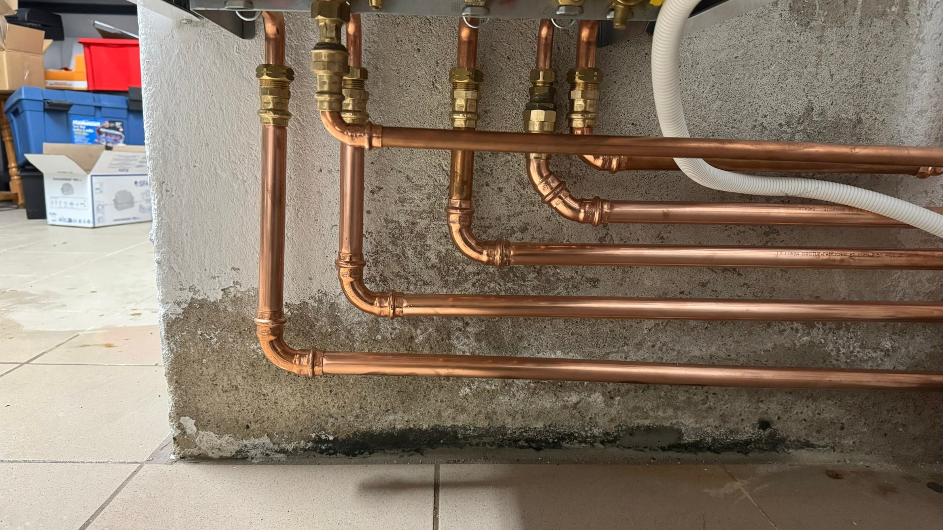 Miedziane rury instalacji grzewczej, z precyzyjnymi złączkami, zamontowane przy szarej, surowej ścianie. Fragment instalacji z białym przewodem. Widoczne skrzynki w tle.