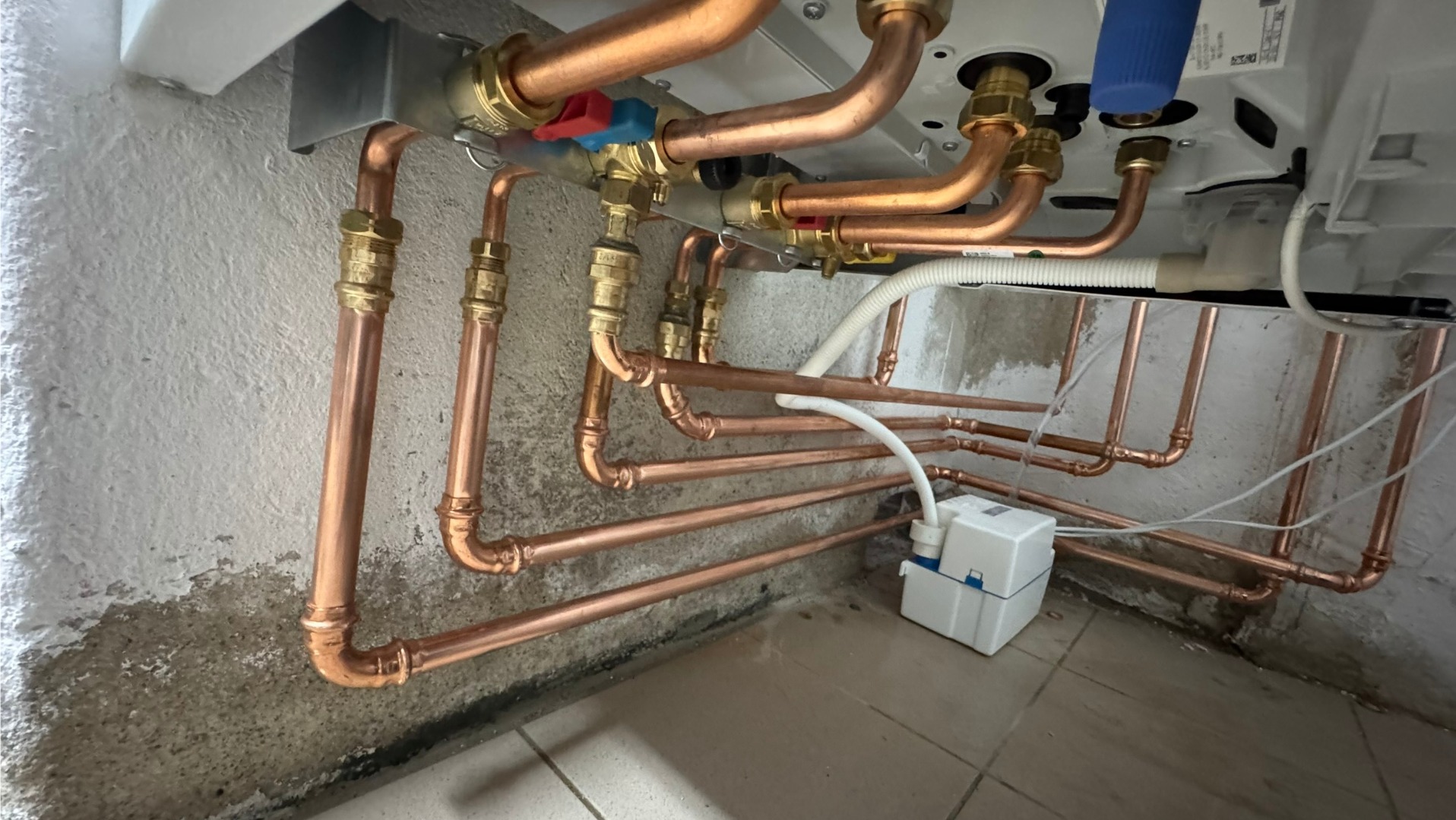 Miedziane rury instalacji grzewczej pod kotłem, z widocznymi zaworami i elementami mocującymi, na tle ściany z surowym wykończeniem i częściowo wykafelkowanej podłogi.