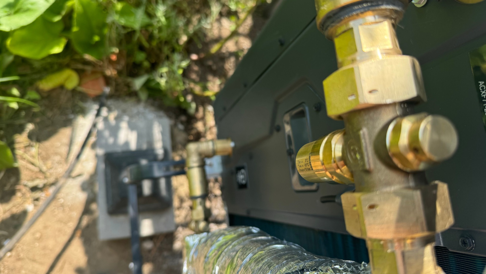 Szczegółowy widok złotych złączek instalacji chłodniczej na tle zieleni, z widoczną rurą w oplocie i fragmentem urządzenia. Kąt ujęcia z dołu podkreśla precyzję montażu.