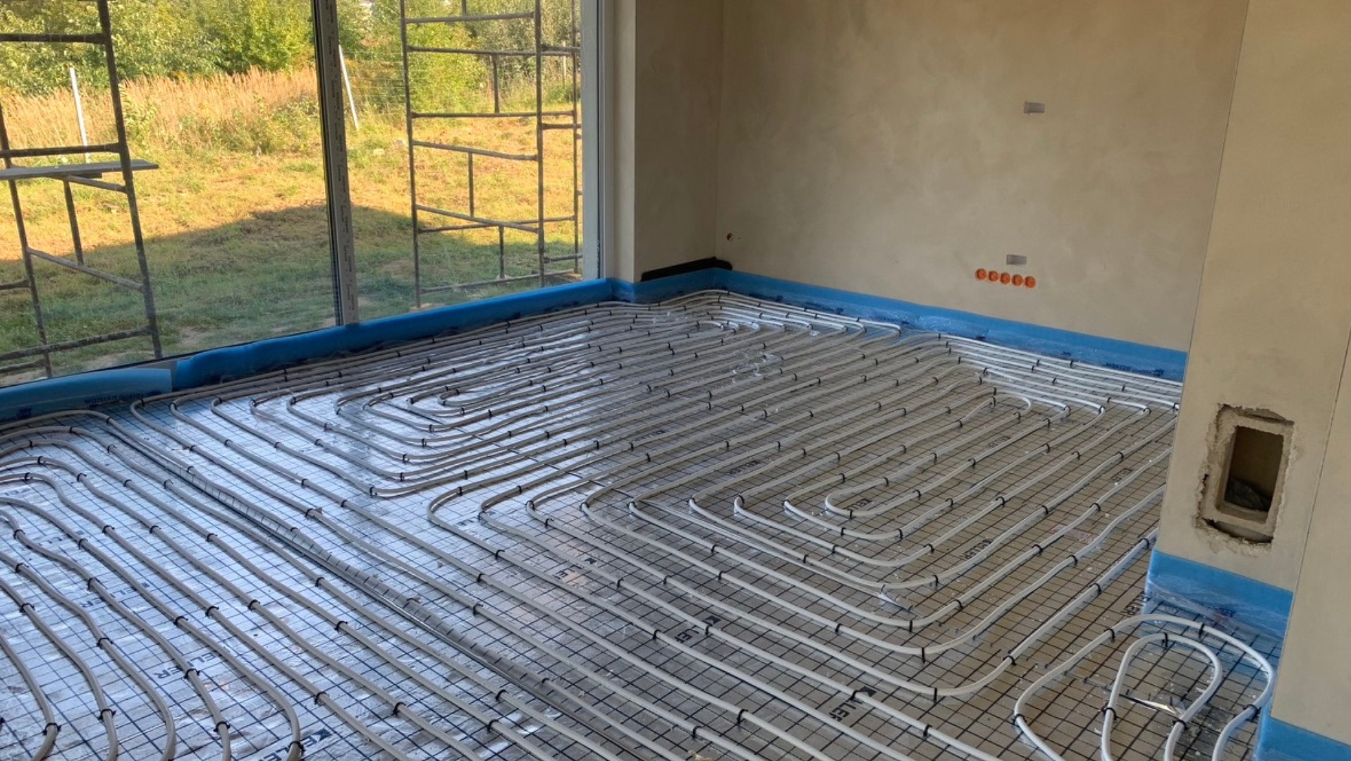Ułożone rury ogrzewania podłogowego na czarnej siatce w jasnym pomieszczeniu z dużym oknem i widokiem na zieleń. Rury zabezpieczone niebieską folią.