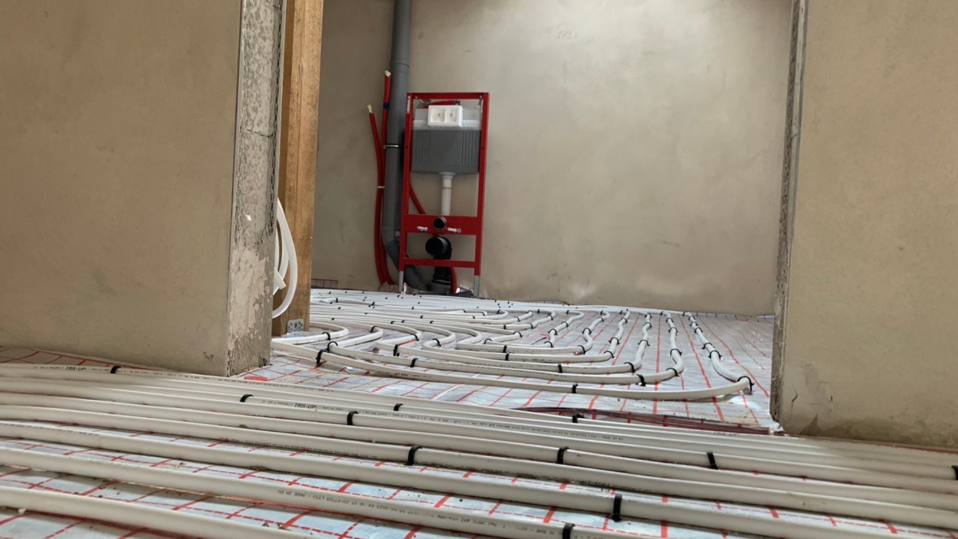 Białe rury ogrzewania podłogowego ułożone na czerwonej macie w surowym wnętrzu, widok przez framugę drzwi, z elementami instalacji wodnej w tle.
