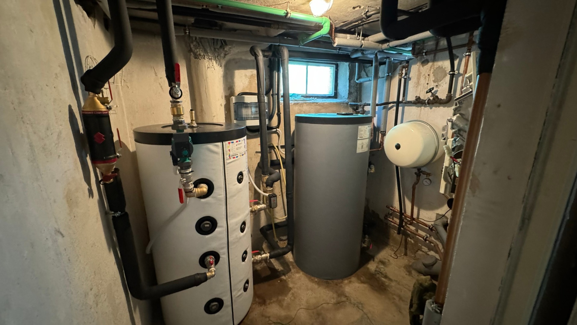 Dwa cylindryczne zbiorniki, biały i szary, w piwnicy z instalacją grzewczą. Widoczne rury, zawory i okno piwniczne. Biały zbiornik z czarnymi okręgami i izolacją.
