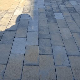 JulJan - Praca brukarza w toku: układanie szarej kostki brukowej. Widoczny cień osoby, młotek i piasek między kostkami. Kąt ujęcia z góry, skupienie na detalu kostki.