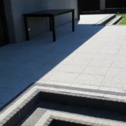 Ułożona kostka brukowa tworząca taras z niskimi schodami, obrzeża z ciemniejszej kostki, stół na tarasie, trawnik w tle, nowoczesny styl.