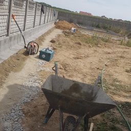 JulJan - Budowa ogrodzenia panelowego z podmurówką prefabrykowaną. Widoczna taczka, piła do betonu i narzędzia. W tle domy i drzewa.