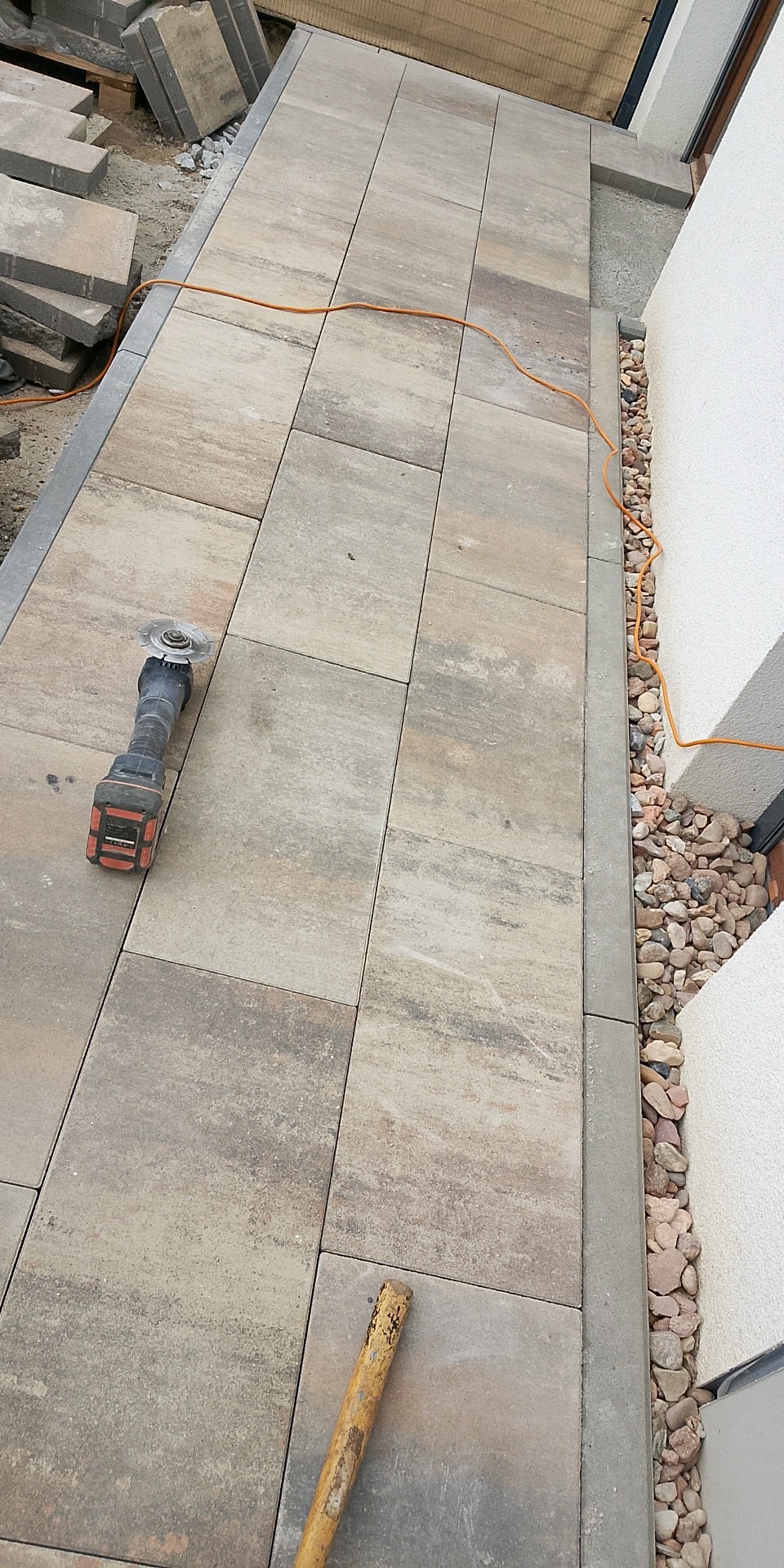 Ułożona, beżowo-szara kostka brukowa wzdłuż budynku. Widoczna szlifierka kątowa, kabel i drewniany trzonek. Obrzeże z małych kamieni.