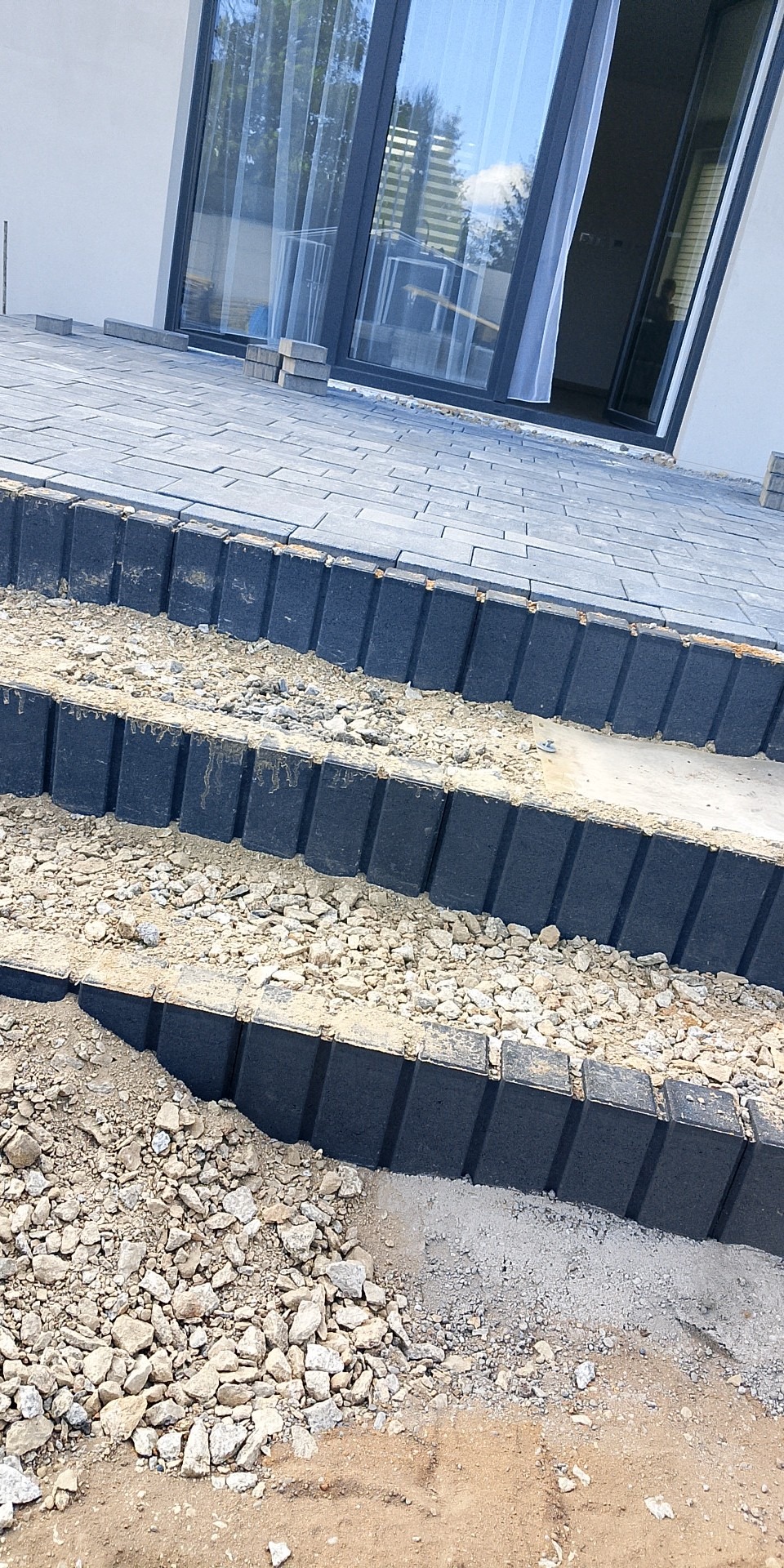 Schody z kostki brukowej w trakcie budowy, z widocznym podsypem żwirowym i częściowo ułożoną nawierzchnią, prowadzące do nowoczesnego budynku z dużym oknem.