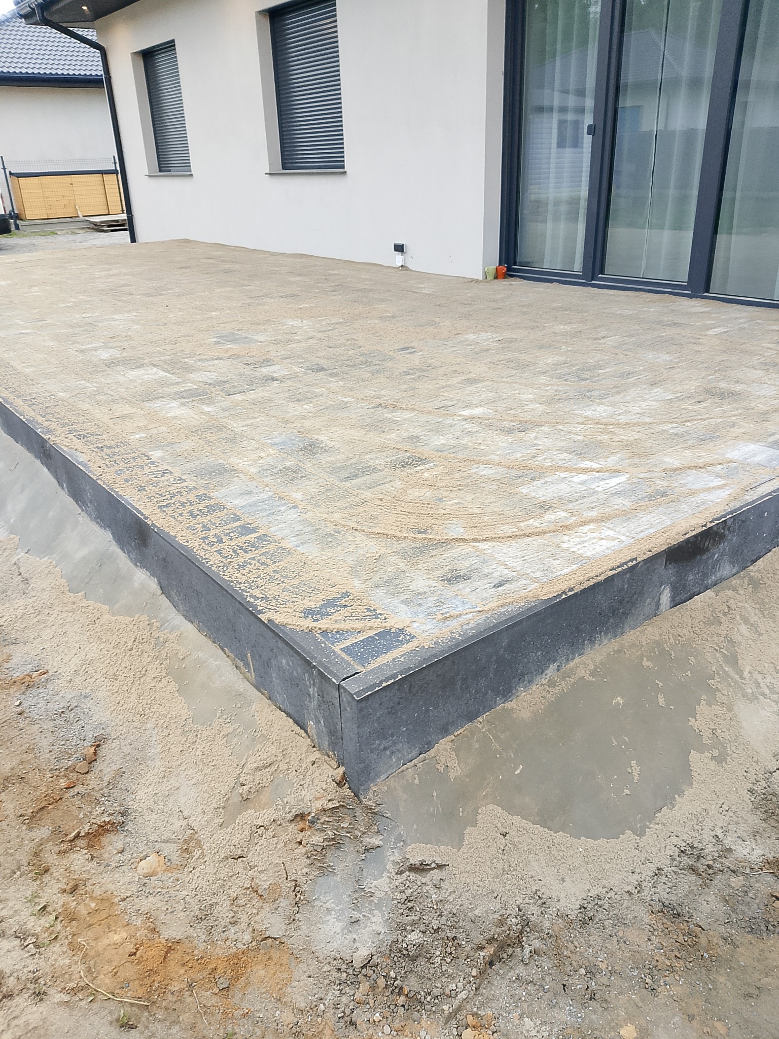 Nowa nawierzchnia z kostki brukowej na posesji, z widocznymi obrzeżami i piaskiem, w trakcie prac wykończeniowych przy domu z szarymi oknami.