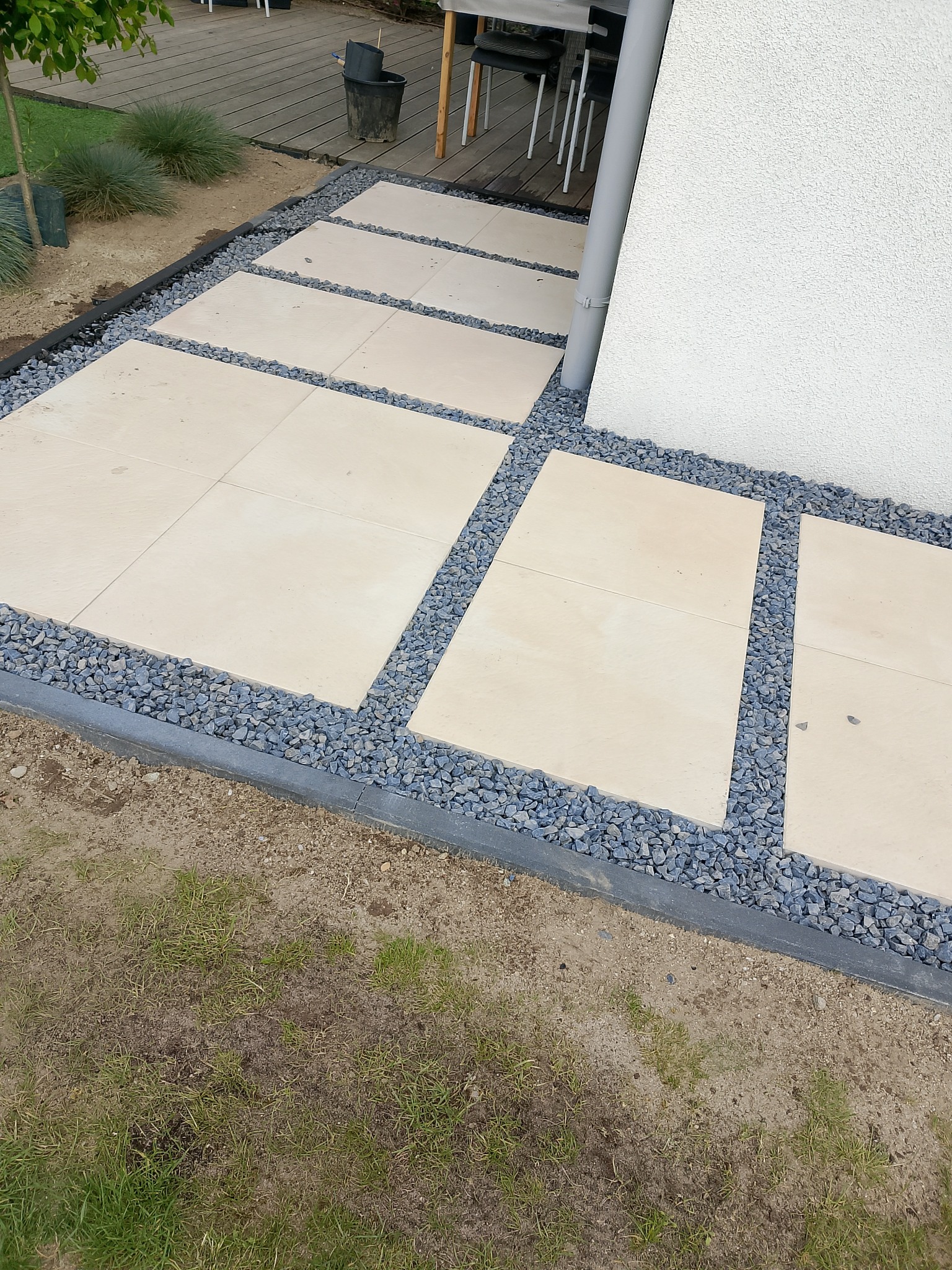 Nowoczesna ścieżka z jasnych płyt, otoczona szarym żwirem i obrzeżem, prowadząca wzdłuż białej ściany budynku. Minimalistyczny design i staranne wykonanie.