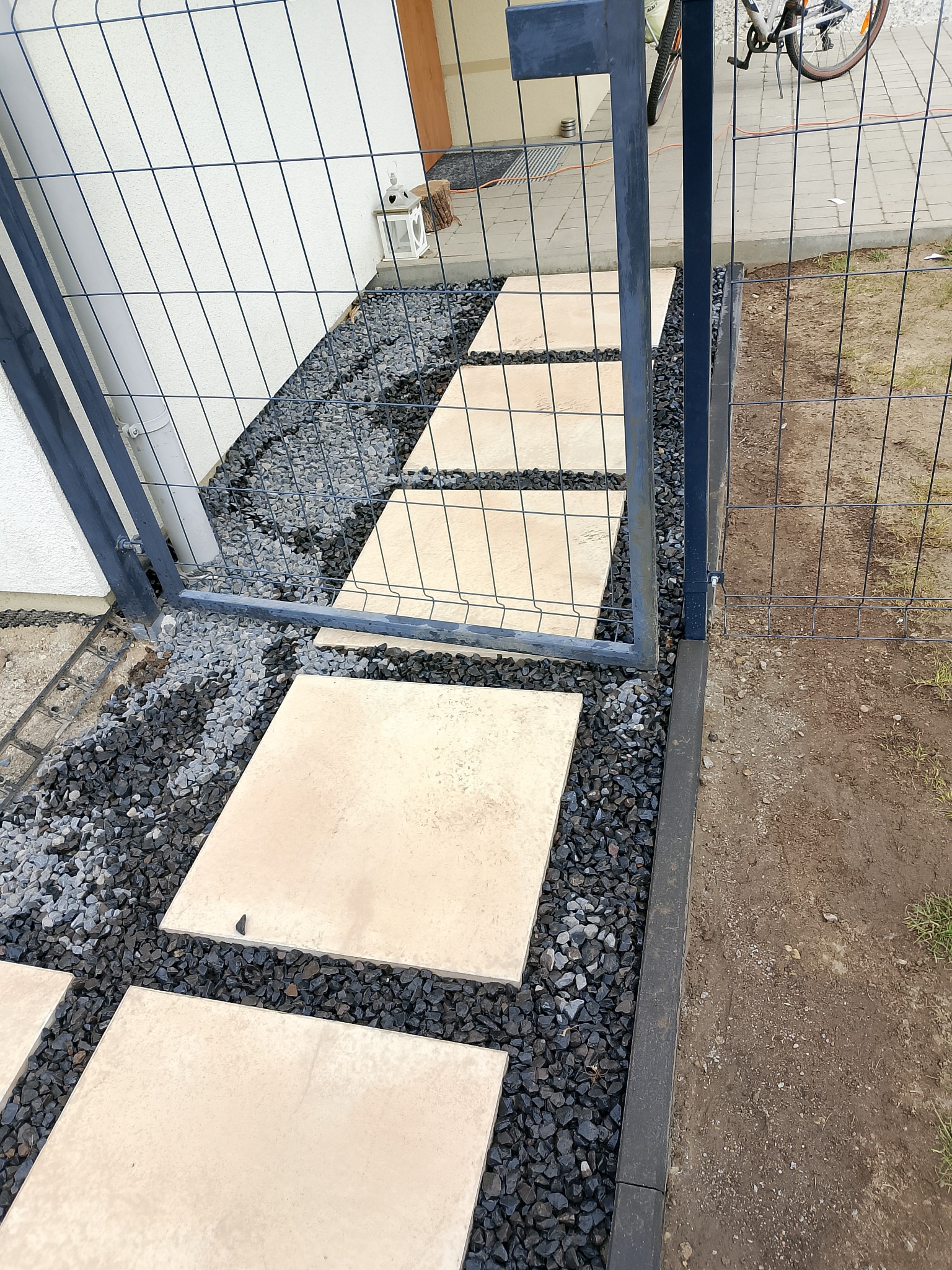 Ścieżka z płyt chodnikowych ułożona w ogrodzie, otoczona czarnym grysem, widoczna brama z siatki, nowoczesny design.