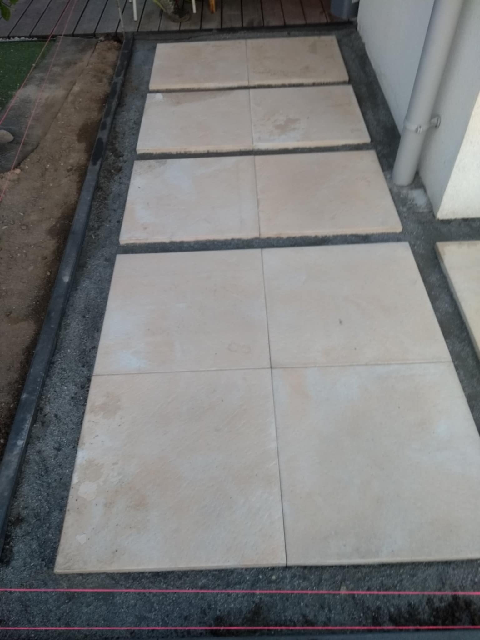 Jasne, kwadratowe płyty ułożone w rzędzie na podsypce, tworzące ścieżkę. Widoczne wytyczone sznurkiem granice, obrzeża i częściowo ułożona nawierzchnia.