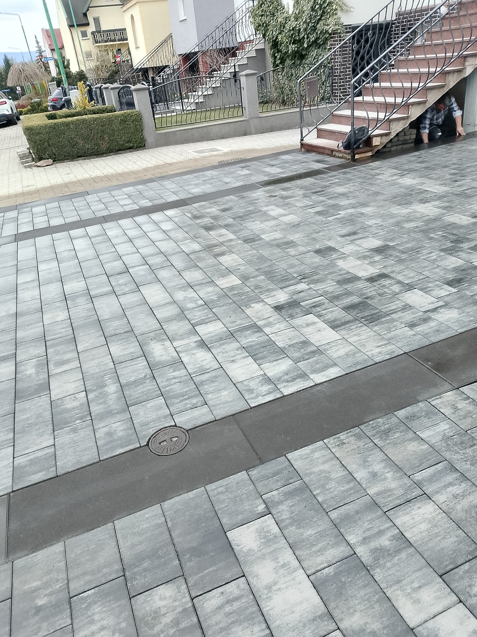 Szara kostka brukowa ułożona w regularny wzór, z ciemniejszym pasem i studzienką kanalizacyjną. Osoba pod schodami budynku w tle.