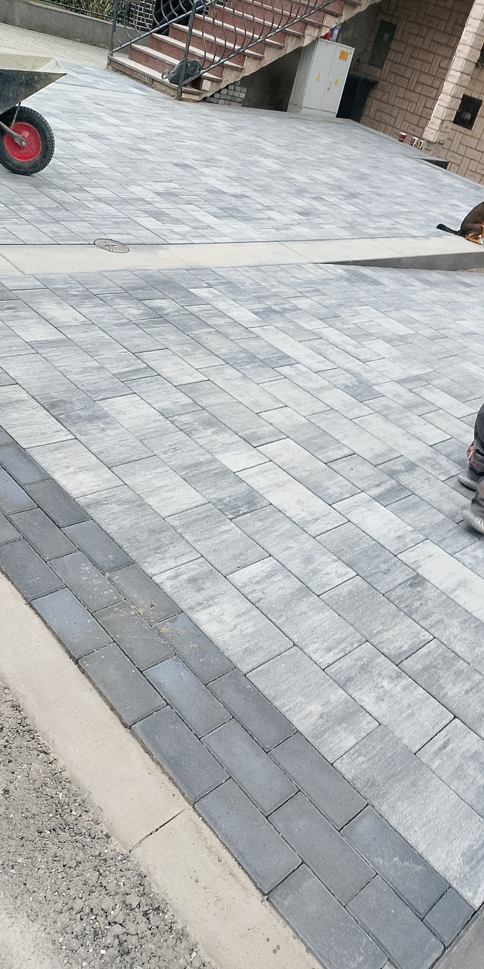 Ułożona szara kostka brukowa na podjeździe, z widocznym fragmentem krawężnika i schodami w tle. W rogu widoczny wózek budowlany.