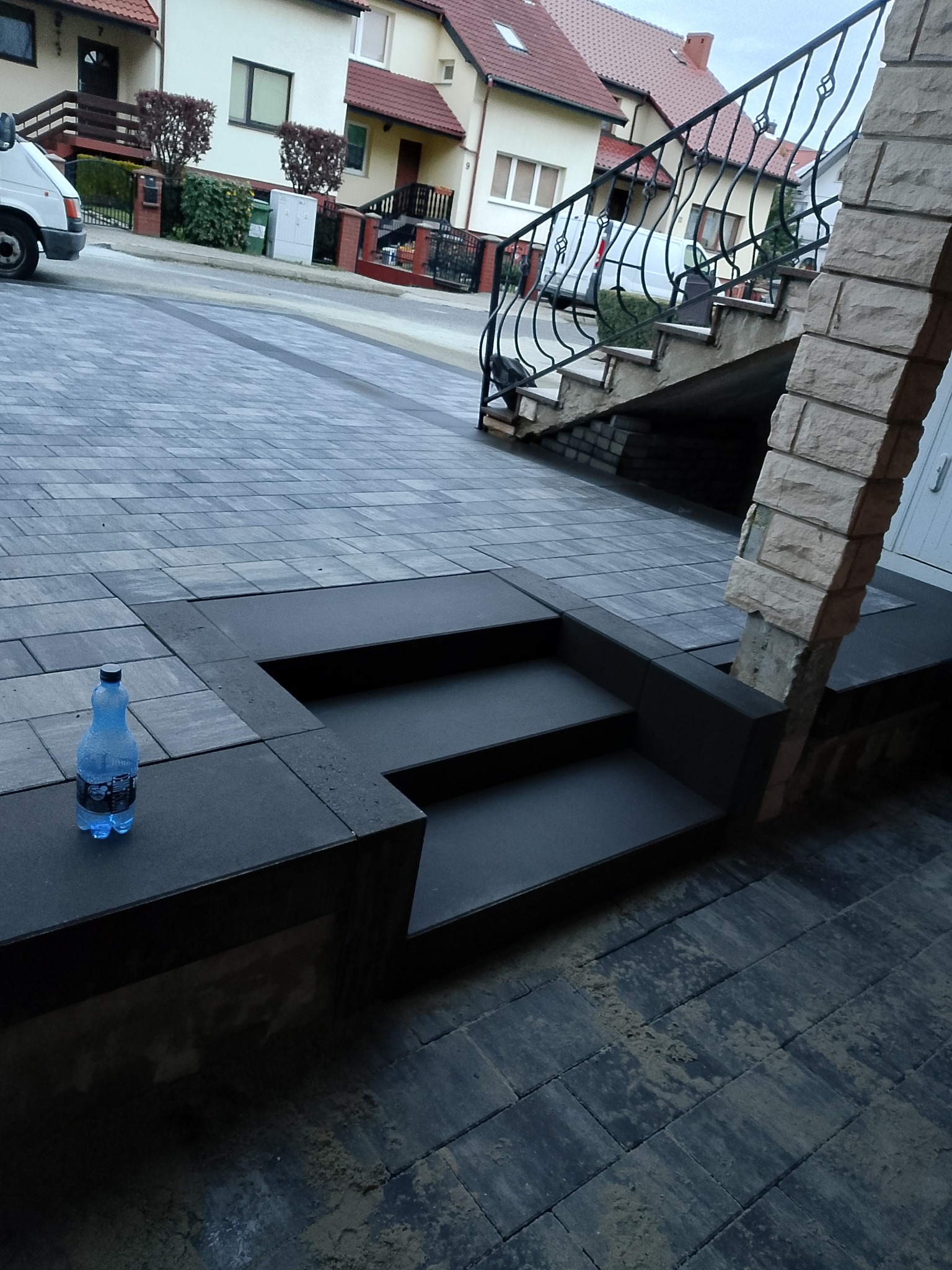Schody z kostki brukowej prowadzące do domu, z butelką wody na krawędzi. Widoczna ulica i zabudowa jednorodzinna w tle. Precyzyjne wykonanie.